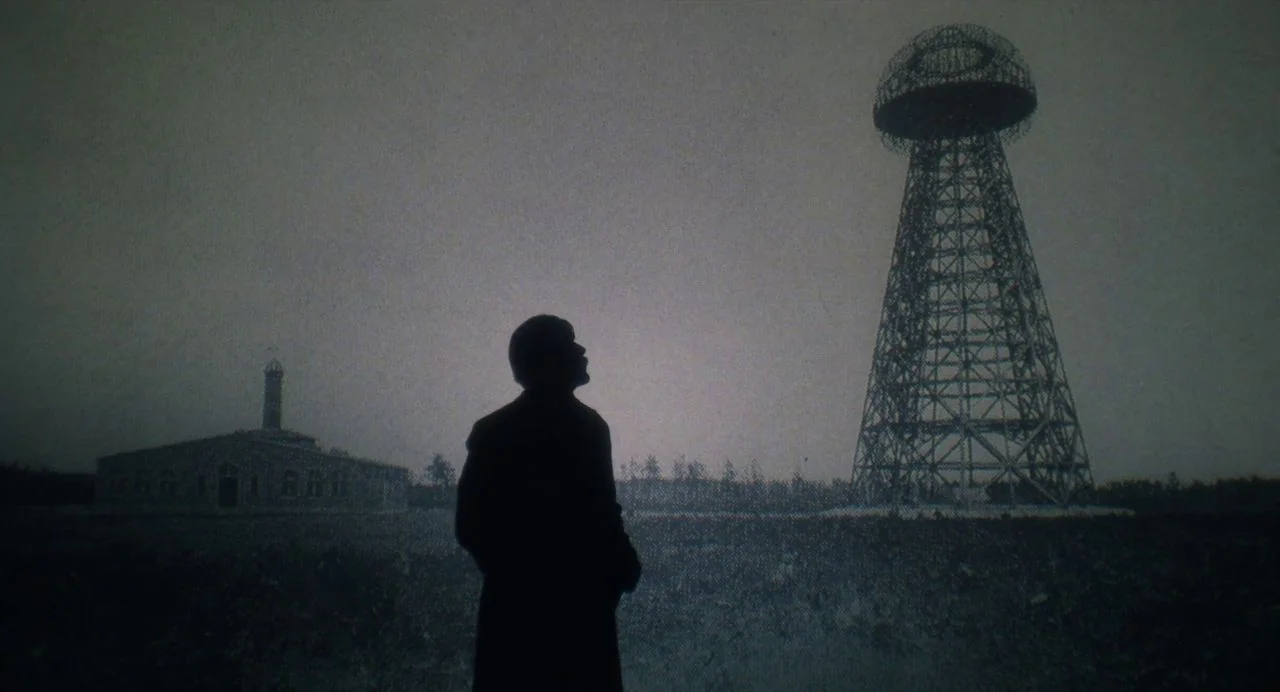Tesla (2020) movie still: wide — A dark field features a tall metal radio transmitter tower against a twilight sky. A lone man stands in the…