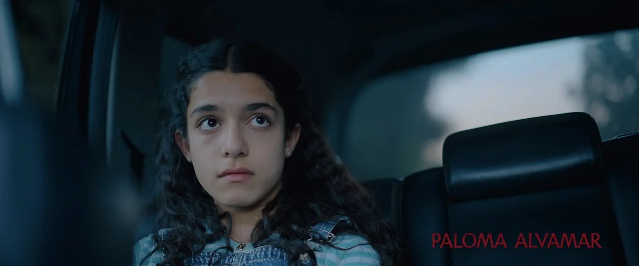 Evil Eye (2020) movie still: medium — A teenage girl sits in the back of a moving car during dusk. She gazes out of the vehicle window while lit by…