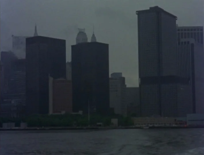 News From Home (1977) movie still: establishing — A view of the lower Manhattan skyline under a heavy, overcast grey sky. Several dark skyscrapers…