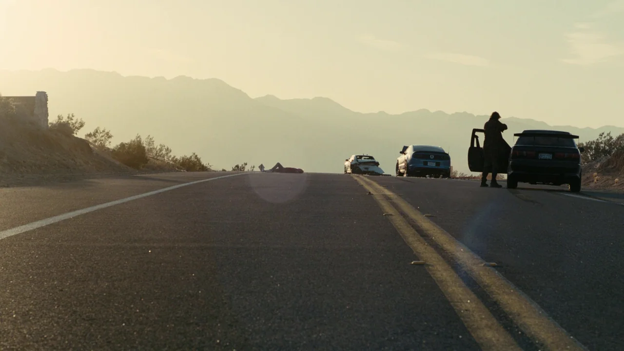 One Battle After Another (2025) movie still: wide — A stretch of paved road in an arid landscape with distant hazy mountains. A man stands near an…