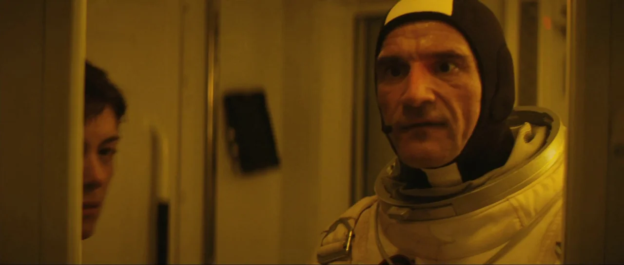 The Last Days On Mars (2013) movie still: medium — A cramped yellow-lit room inside a spacecraft contains a man wearing a space suit. The man gazes…