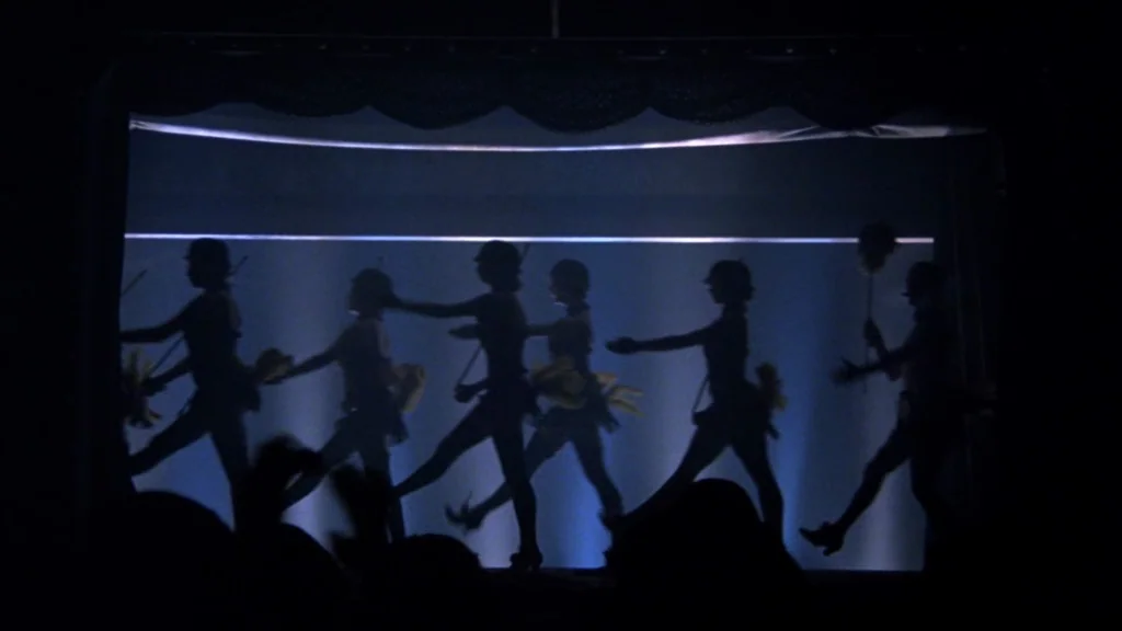 Cabaret (1972) movie still: wide — A dark theater features a backlit screen showing the silhouettes of six dancers. The dancers move in a line…
