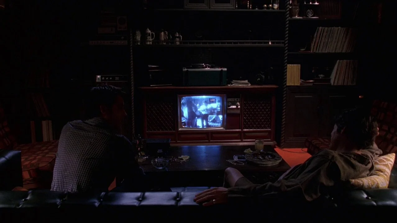 Auto Focus movie still: wide — A dimly lit room features a large wood-paneled television console and built-in shelving units. Two men sit on a black…