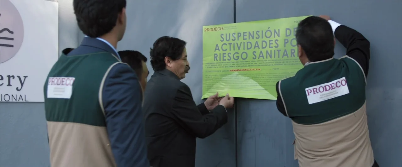 Winner Takes the Cake (2025) movie still: medium — A gray metal wall features a bright green official suspension notice posted by authorities. Three…