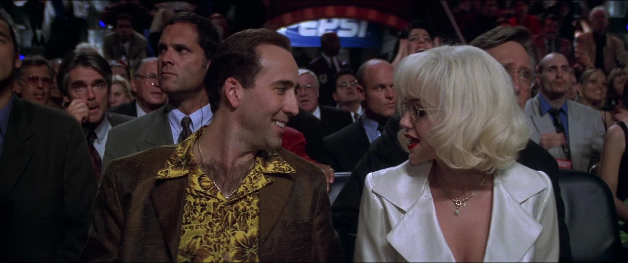 Snake Eyes (1998) movie still: medium — A crowded arena with rows of seats occupied by well-dressed people attending an event. A man and a blonde…
