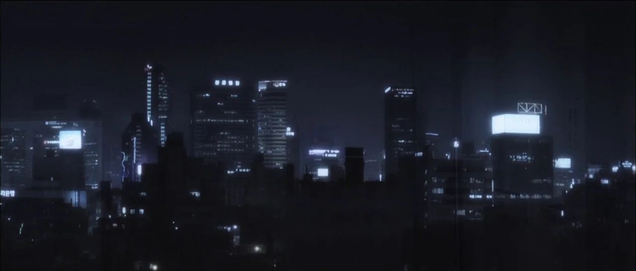 The Chaser (2008) movie still: establishing — A dense urban skyline at night features illuminated skyscrapers with glowing advertisements. The…