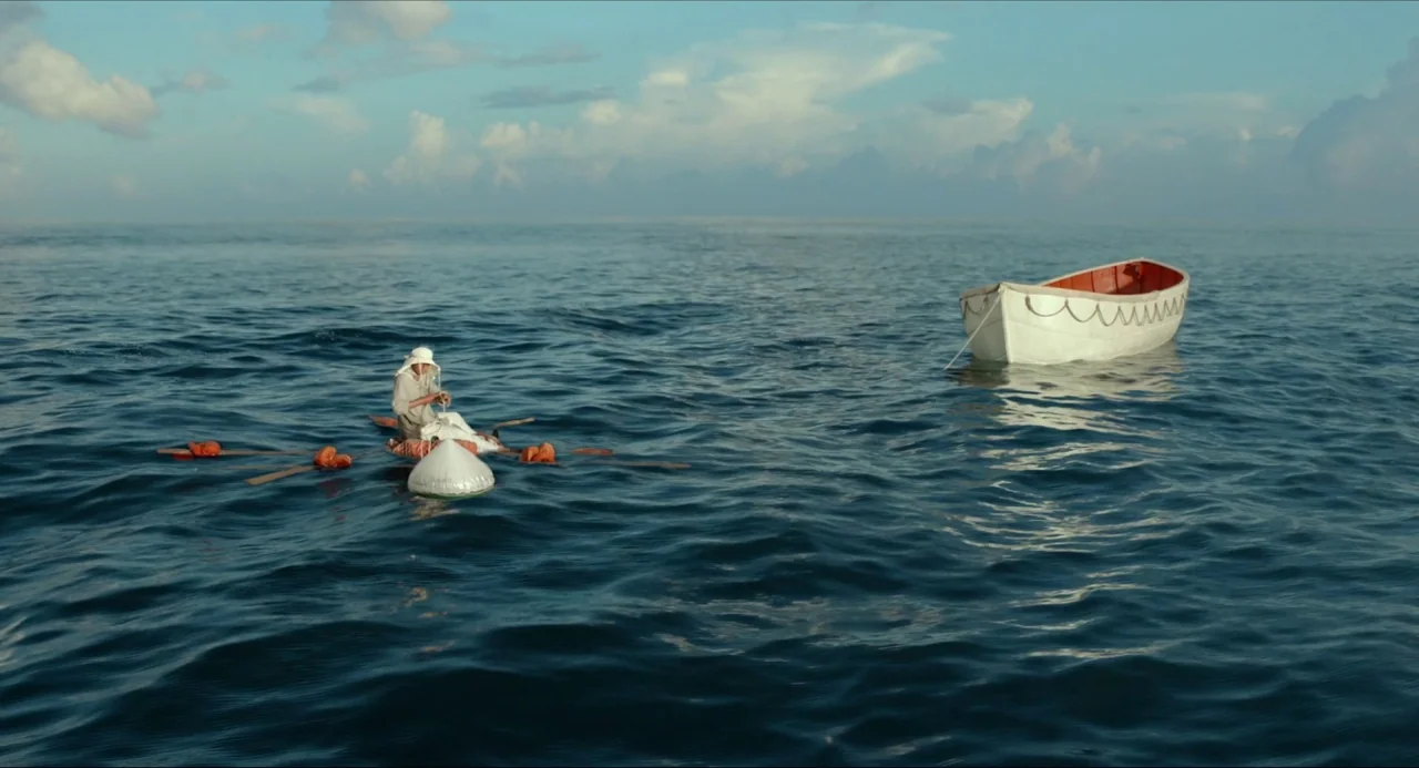 Life of Pi (2012) movie still: wide — The open sea stretches to a horizon line under a pale blue, cloudy sky. A man sits on a small raft near a…
