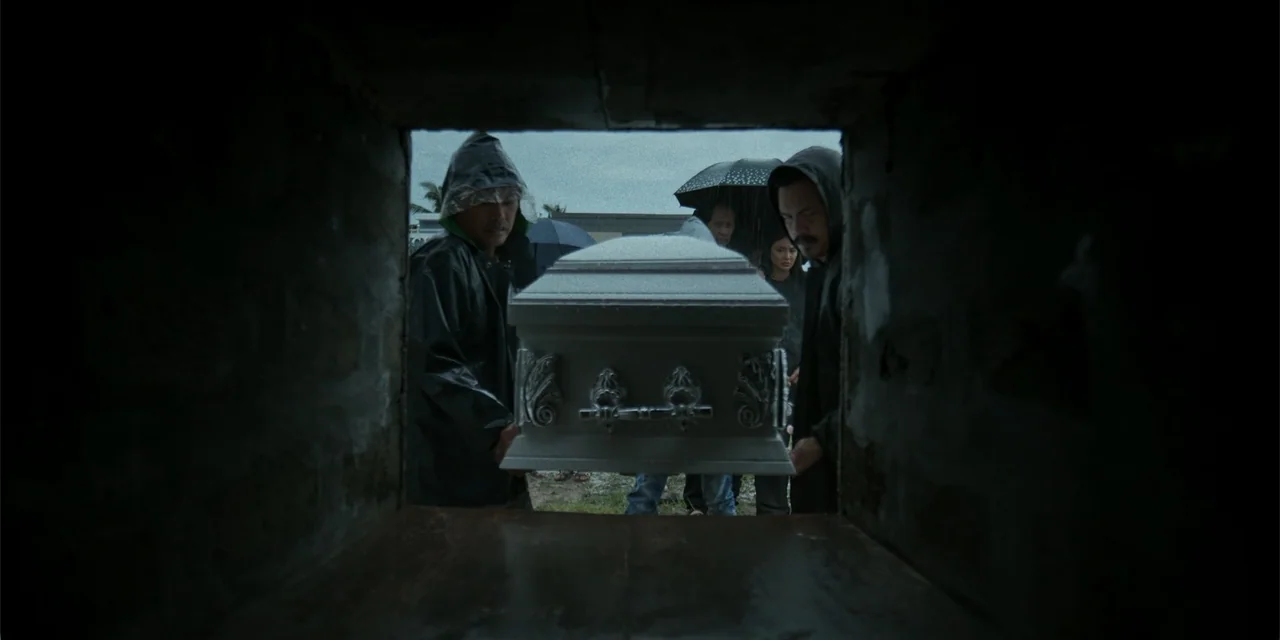 Nokturno (2024) movie still: medium — A group of people under an overcast sky stand around an open grave site. Two men in raincoats hold the silver…