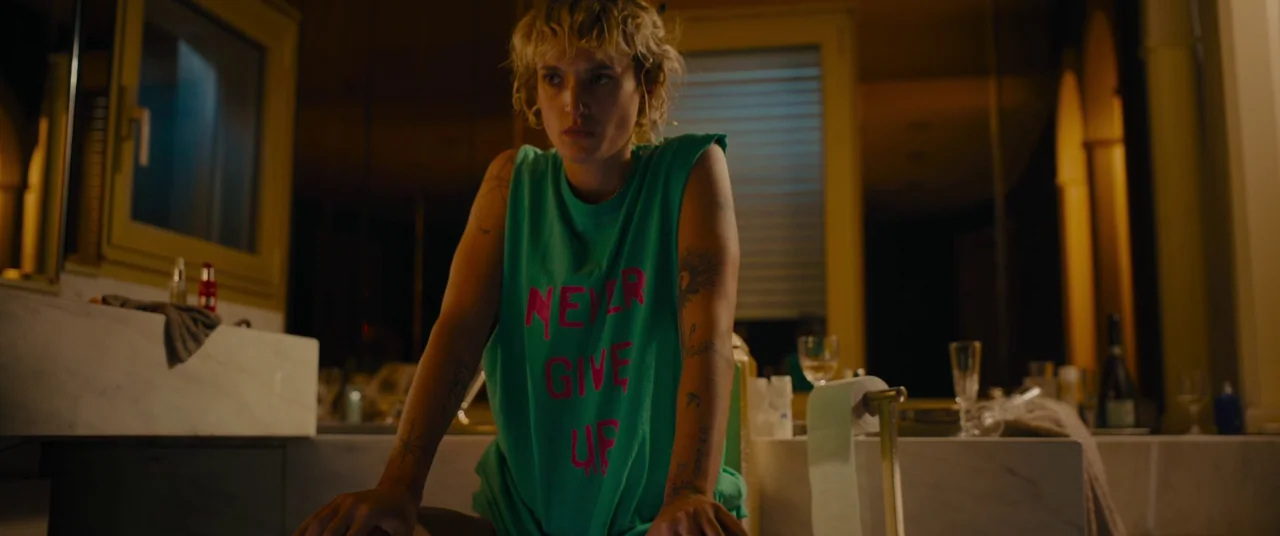 Titane (2021) movie still: medium — A dimly lit room features a marble counter and metallic fixtures under warm lighting. A woman with short blonde…