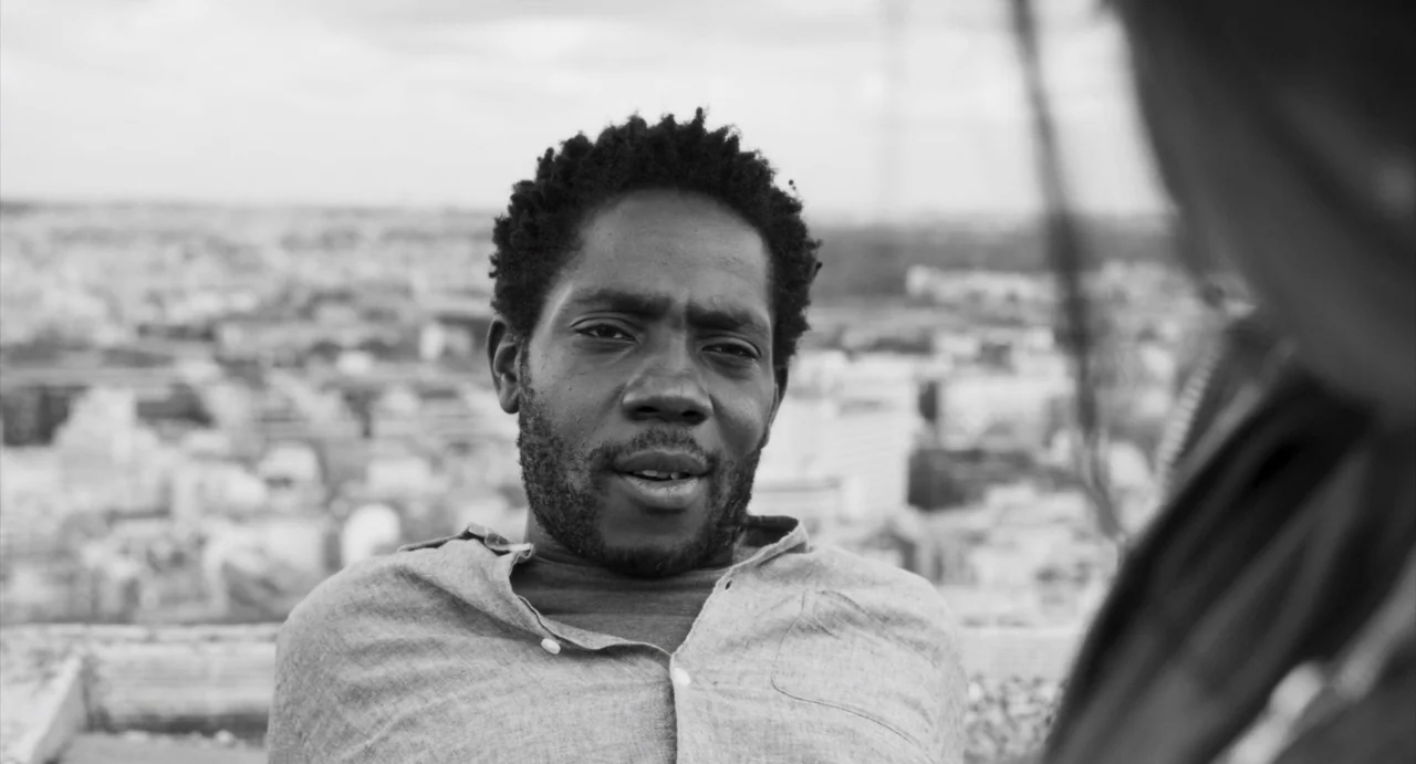 Paris, 13th District (2021) movie still: medium — A man sits on a rooftop with an out-of-focus cityscape in the background. The man gazes toward…