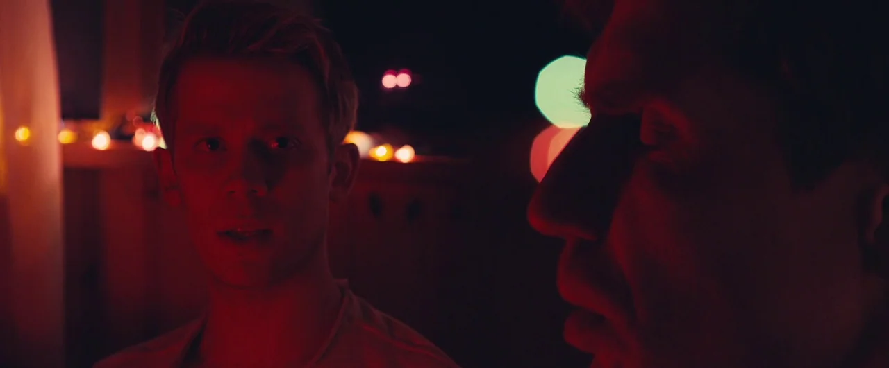 Rift (2017) movie still: medium — A dark indoor space is lit by red light and distant blurry holiday lights. Two men stand in the shadows, one…