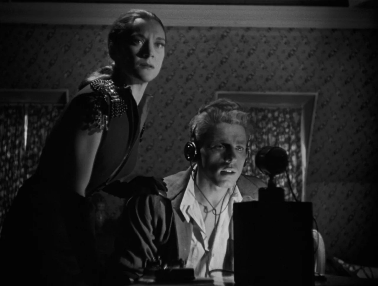 Orpheus (1950) movie still: medium — A dimly lit room with patterned wallpaper contains a radio broadcast setup. A man wearing headphones speaks…