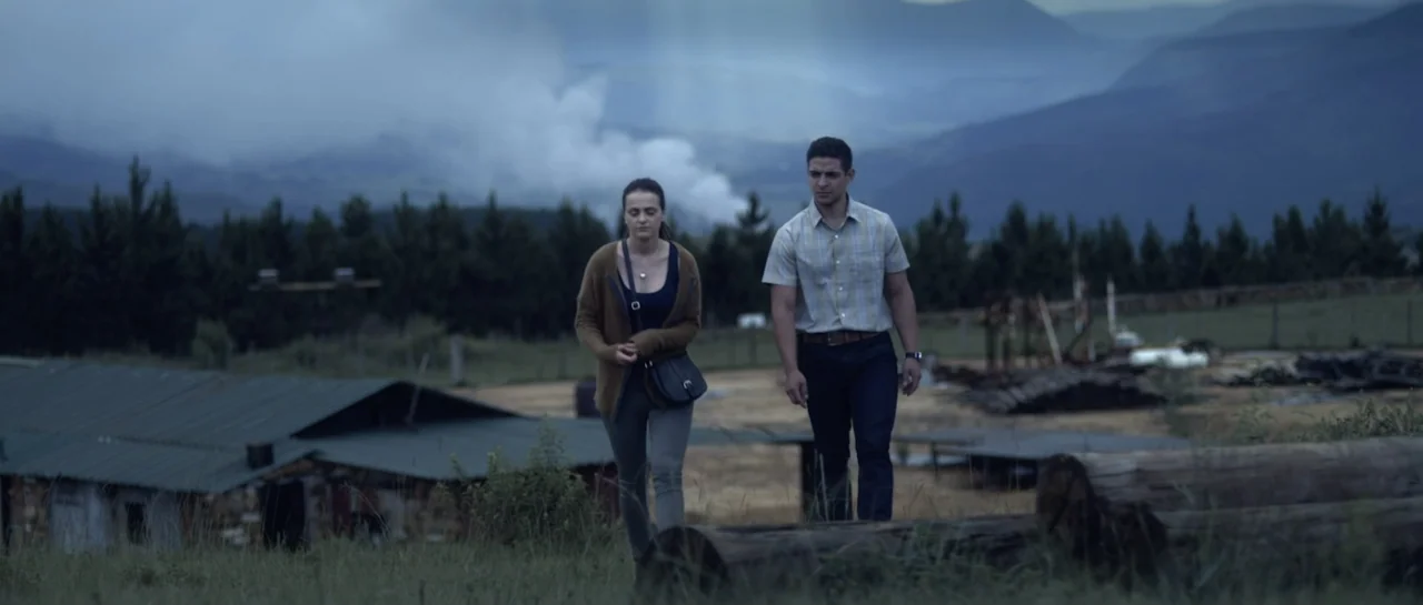The Lullaby (2019) movie still: fullbody — A rural expanse features a grassy foreground, a wooden shed, and distant blue mountains. A man and a…