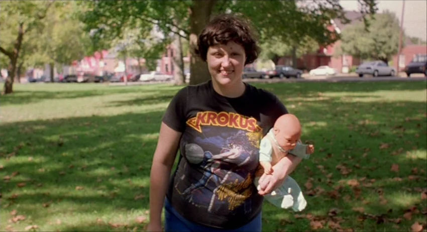 Gummo (1997) movie still: medium — A woman stands in a public park during the day with trees and houses visible. She holds a small plastic baby doll…