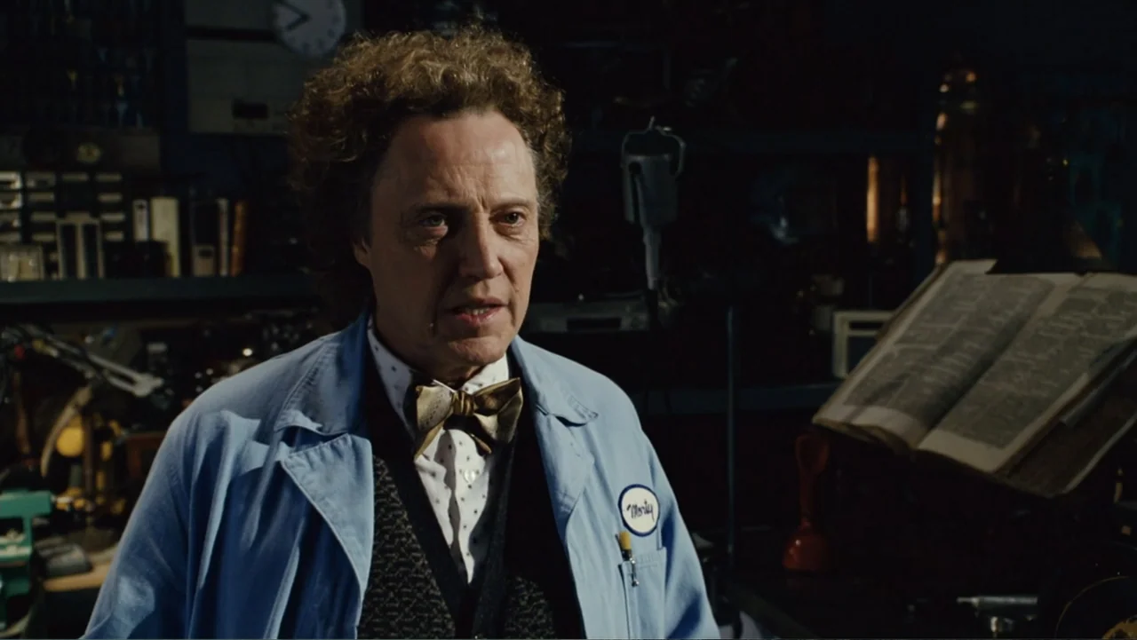 Click (2006) movie still: medium — A cluttered workshop interior with shelves, tools, and an open book. A man wearing a lab coat and a bow tie…