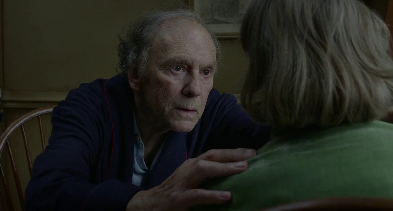 Amour (2012) movie still: medium — An interior dining room with dark wood walls and a framed artwork in shadow. An elderly man leans forward to…