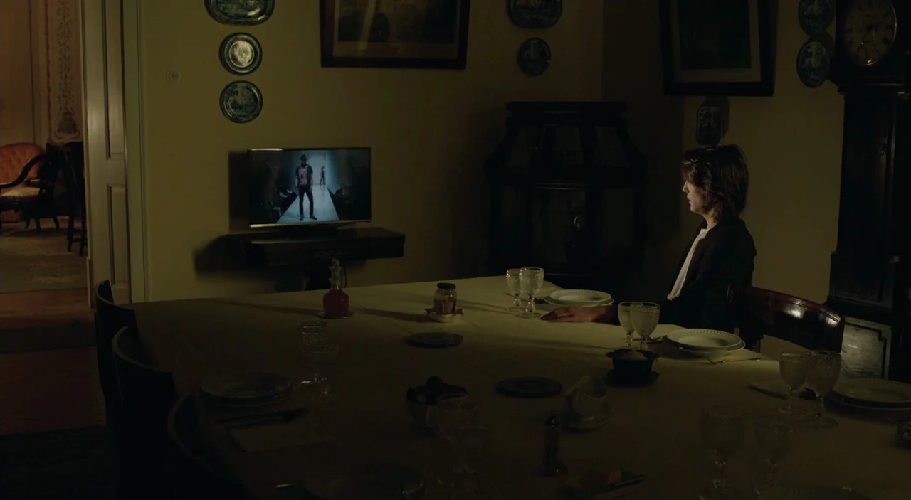 Cosmos (2015) movie still: wide — A dimly lit room features a large table set for a meal in the foreground. A young woman sits at the far end of the…