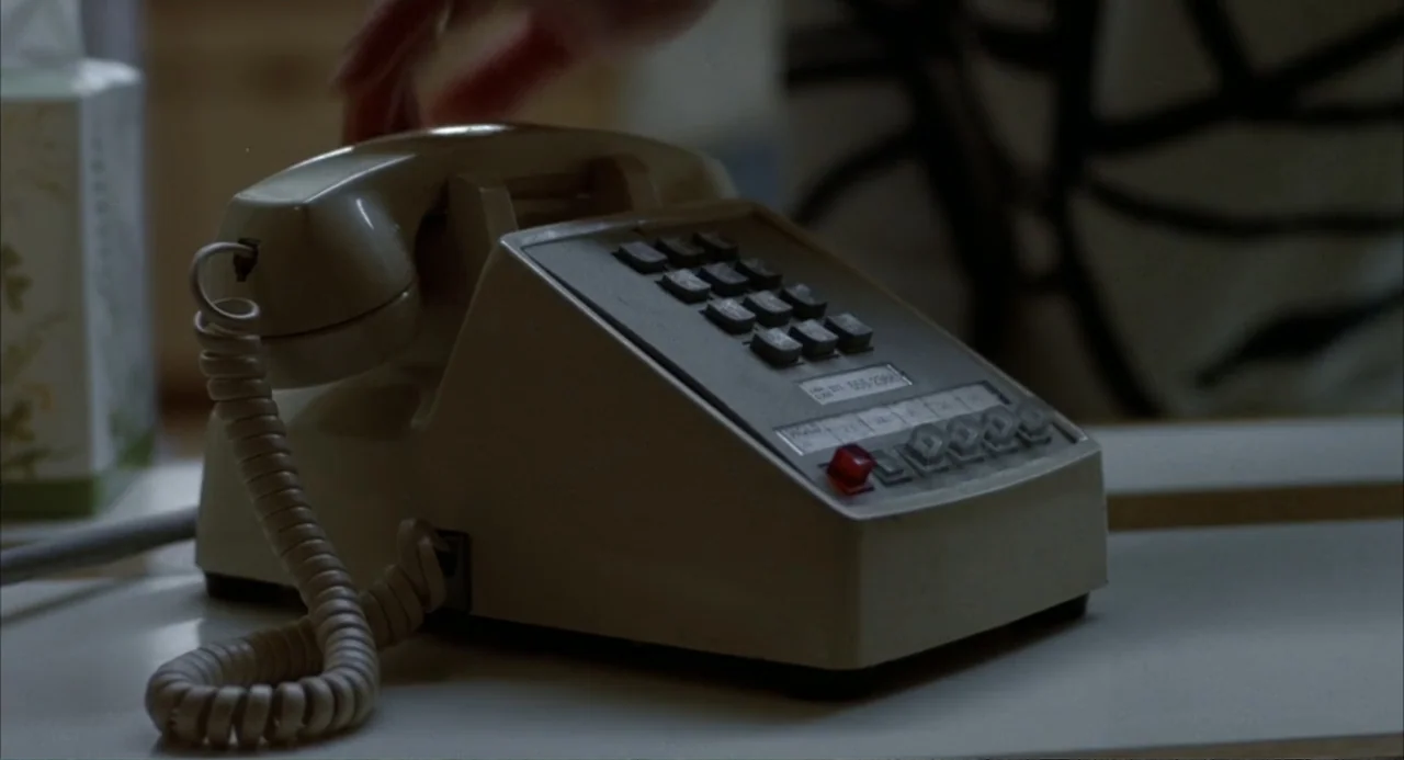 Poltergeist III (1988) movie still: closeup — A beige multi-line business telephone sits on a white desk surface. A hand enters from the top edge to…