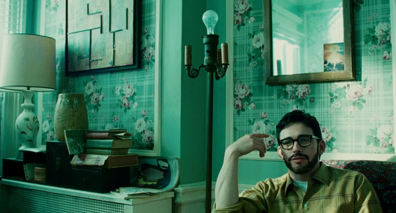 Howl (2010) movie still: medium — A living room with vintage floral wallpaper and a cluttered shelf of books. A man sits in front of the wall and…