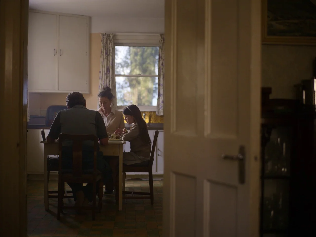 The Quiet Girl (2022) movie still: medium — A 1970s interior with off-white cabinetry and tile floors seen through a doorway. An adult male, an…