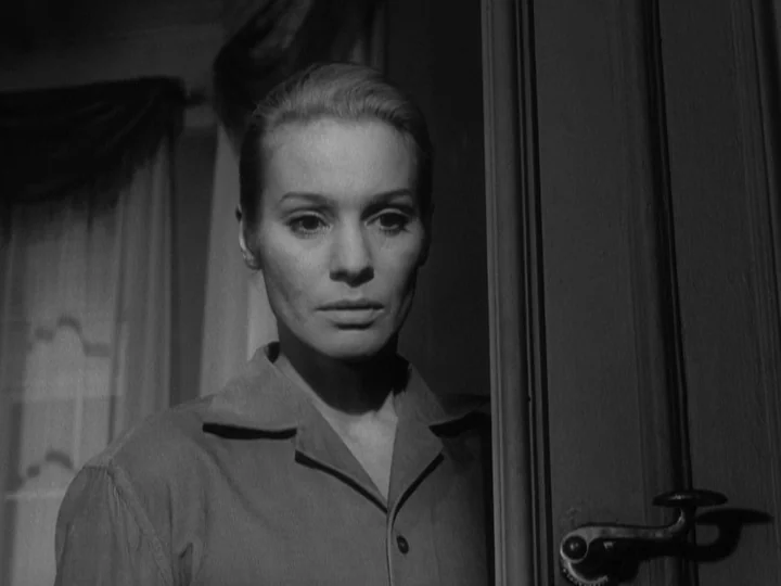The Silence (1963) movie still: medium — A woman stands in a doorway against a backdrop of draped curtains. She faces forward while positioned…