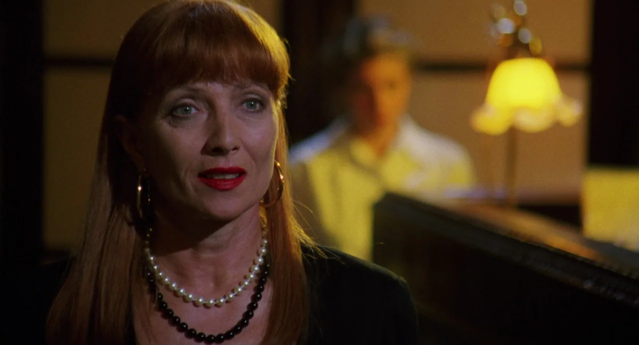 The Spider Labyrinth (1988) movie still: closeup — A dimly lit room contains a reception desk with a yellow glowing lamp. A woman with red hair…