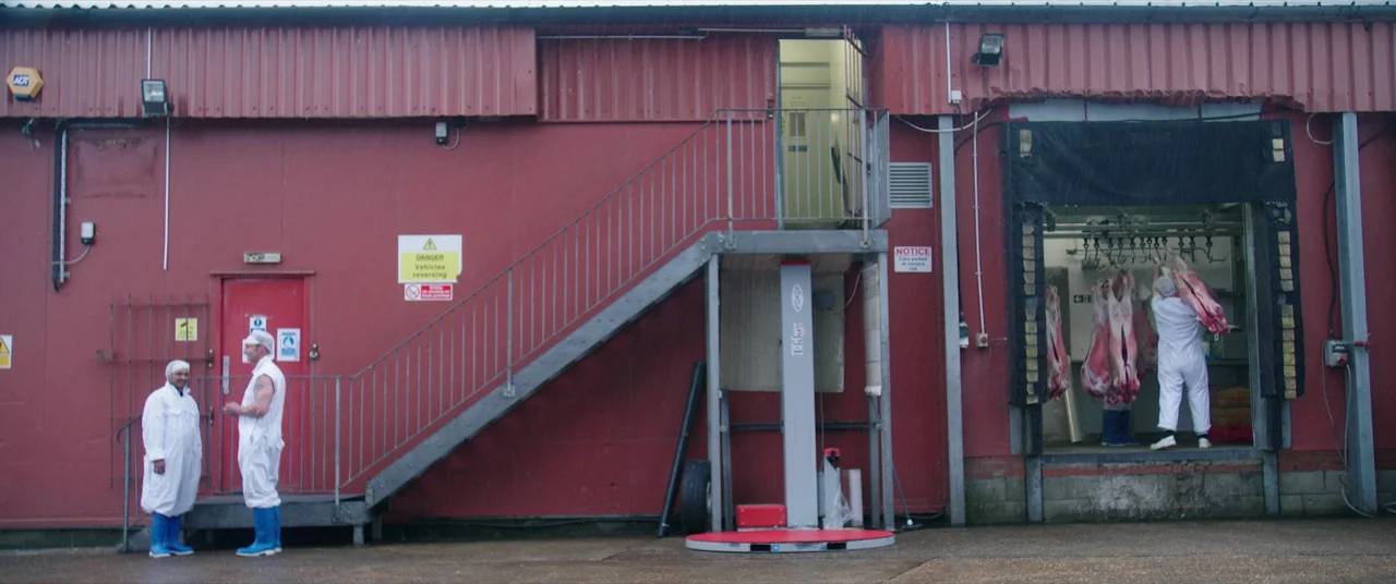 Bull (2021) movie still: wide — The exterior wall of a red industrial building features a staircase and loading bay. Workers in white protective…