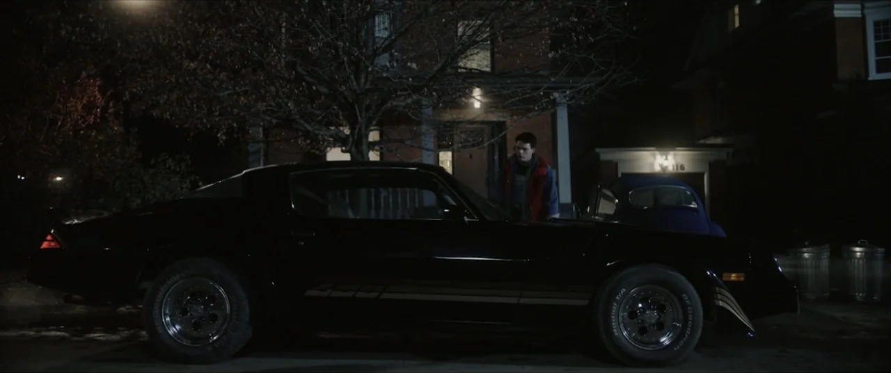 Vicious Fun (2021) movie still: wide — A residential suburban street at night features a dark house and large bare trees. A young man walks past a…