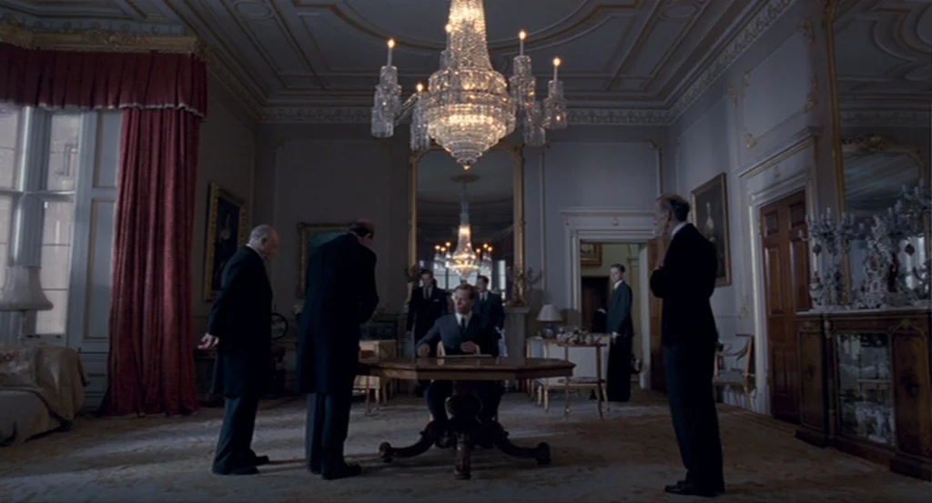 The King’s Speech (2010) movie still: wide — A high-ceilinged room features ornate wall molding, heavy red drapes, and a central chandelier. Several…
