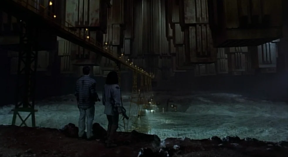 Total Recall (1990) movie still: wide — A dark industrial landscape features massive overhanging structures and a long metal gantry bridge. Two…