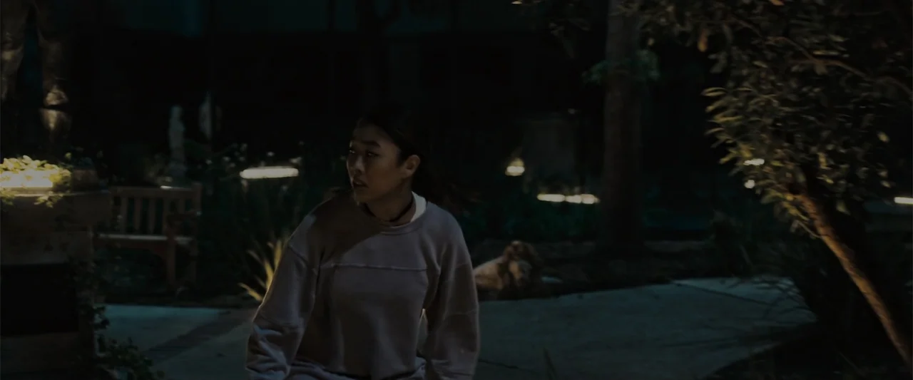 Init!ation (2021) movie still: medium — A dark outdoor park at night features low-level path lighting and thick surrounding vegetation. A young…