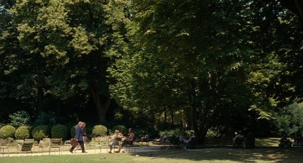 Things to Come (2016) movie still: wide — A sunlit park area features a paved walkway lined with numerous metal chairs. Several individuals are…