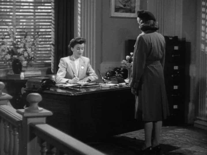 The Seventh Victim (1943) movie still: medium — A 1940s office with a large dark wooden desk and vertical window blinds. A woman sits behind the…