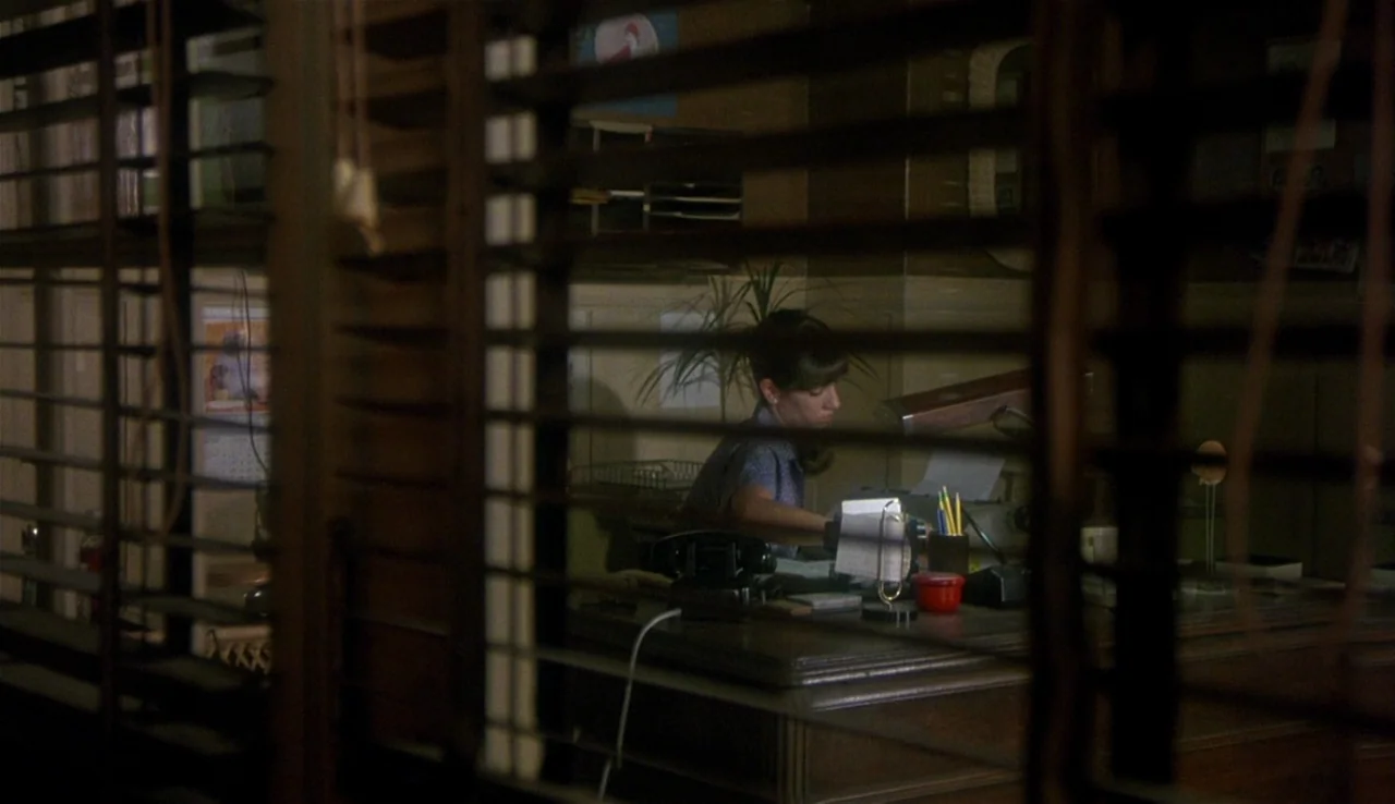 Body Heat (1981) movie still: medium — A dimly lit room with a wooden desk seen through horizontal window blinds. A woman works at a typewriter in…