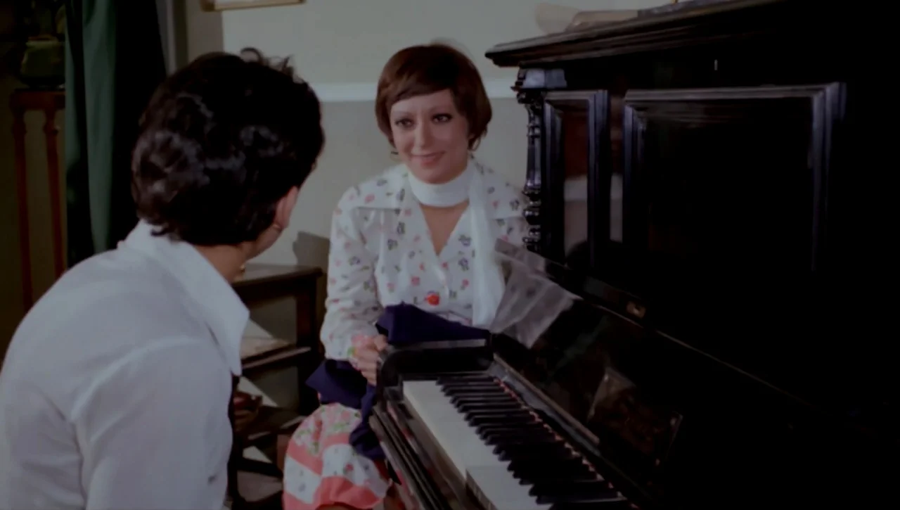 Alexandria…Why? (1979) movie still: medium — A man and a woman interact near an upright piano in a domestic setting. The woman sits at the piano…