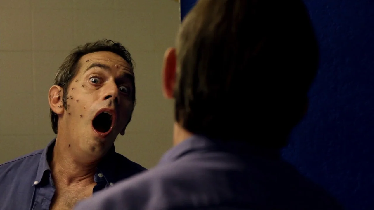 The ABCs of Death (2012) movie still: medium — A man looks into a bathroom mirror with a blue wall reflected behind him. Small insects crawl across…