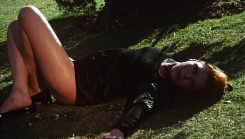 A Bay of Blood (1971) movie still: fullbody — A woman lies on a sloping grass surface outdoors in bright, direct sunlight. She is positioned on her…