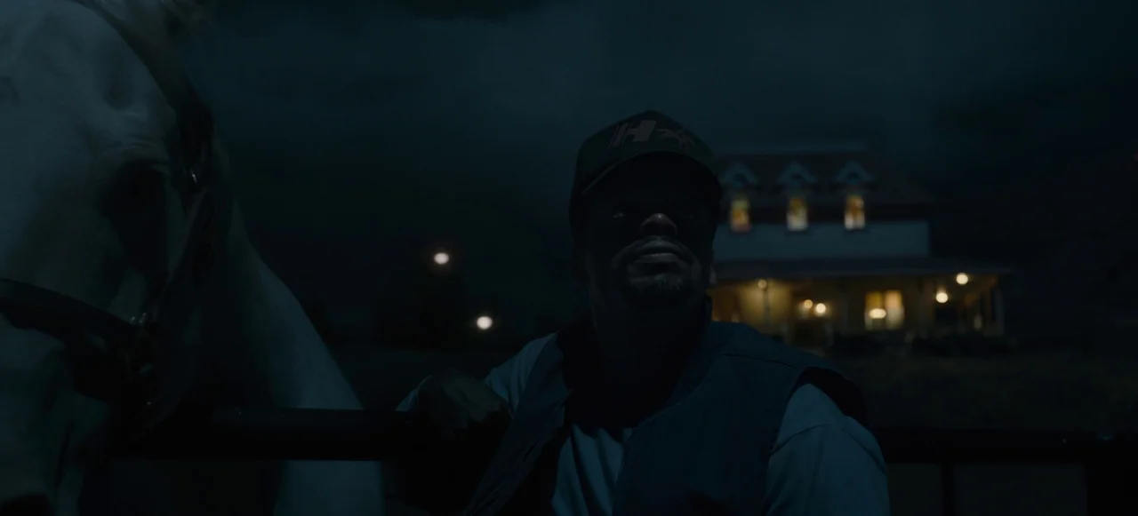 Nope (2022) movie still: medium — A man stands outside at night near a horse, with a distant illuminated house visible. He gazes upward into the…