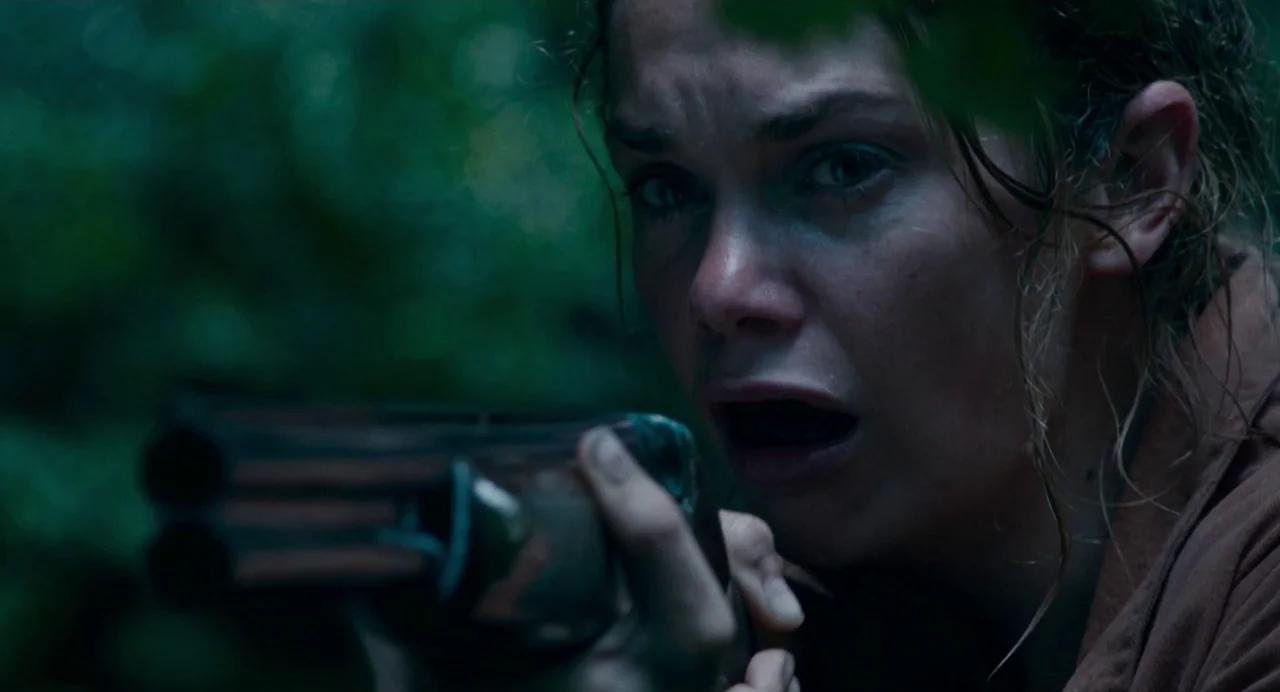 Dark River (2017) movie still: closeup — A woman stands in dense woods with foliage obscuring the upper frame. She holds a shotgun forward while…