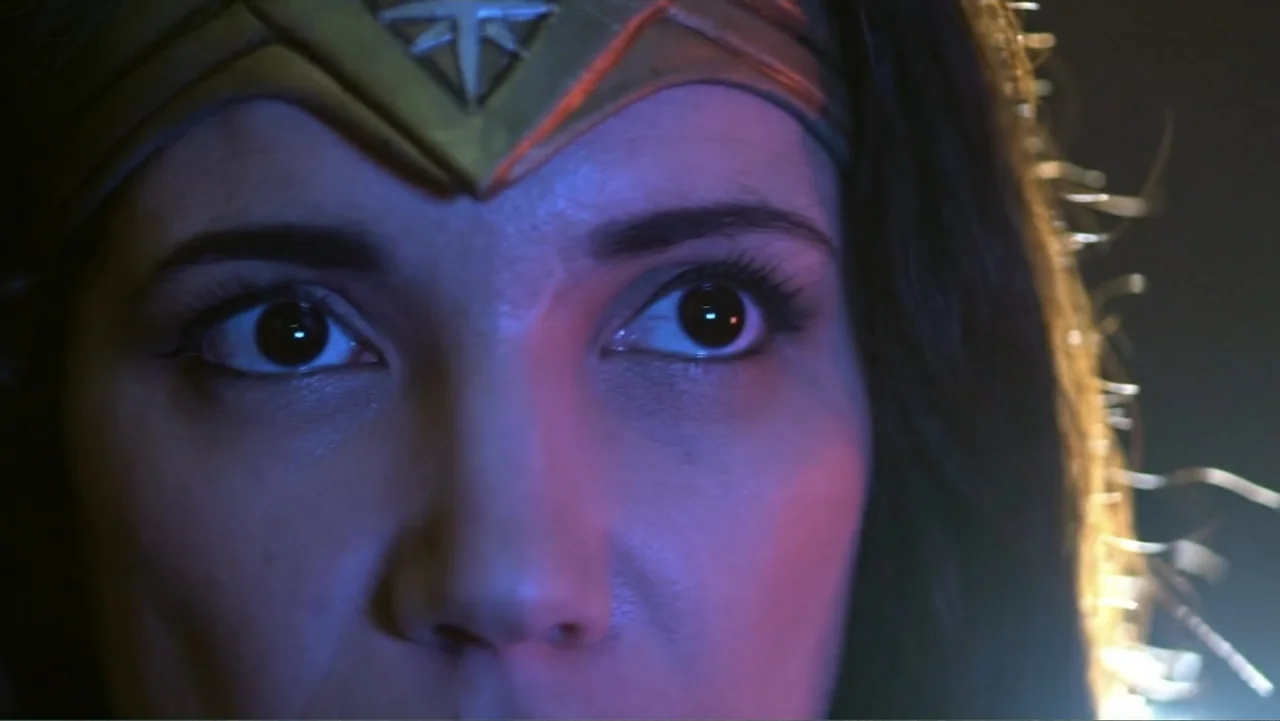 Reign of the Superwomen (2021) movie still: closeup — A close-up view of a woman wearing a metallic headpiece in a dark space. The subject gazes…