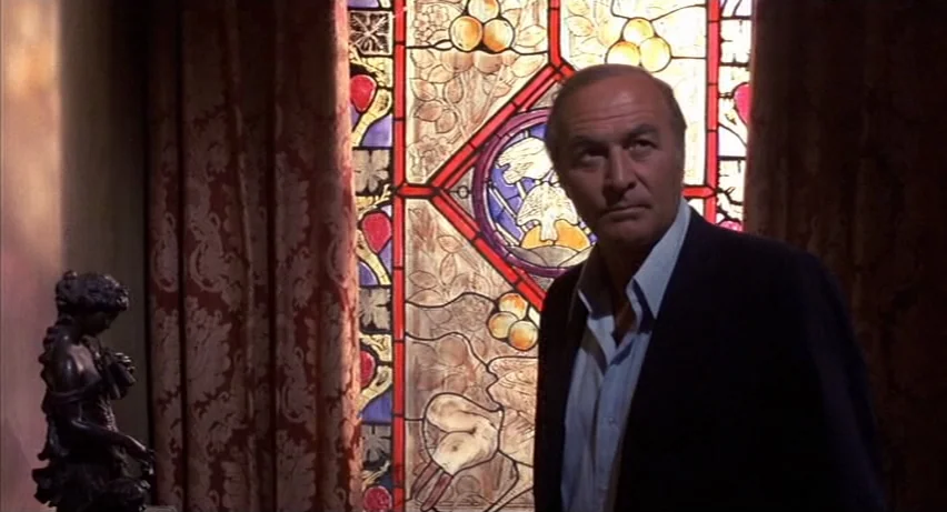 Psycho 2 (1983) movie still: medium — An indoor space features heavy patterned curtains and a large stained glass window. A middle-aged man stands…