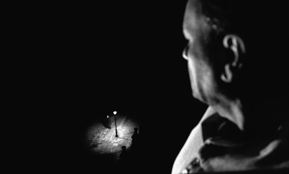 The Man From London (2007) movie still: closeup — A dark night exterior features a cobblestone surface illuminated by a single streetlamp. A man in…