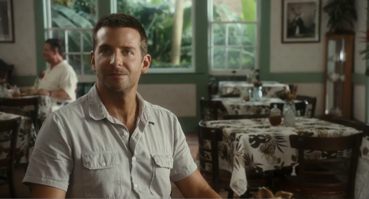 Aloha (2015) movie still: medium — A quiet dining area features tables with floral tablecloths and view of green foliage. A man sits at a table…