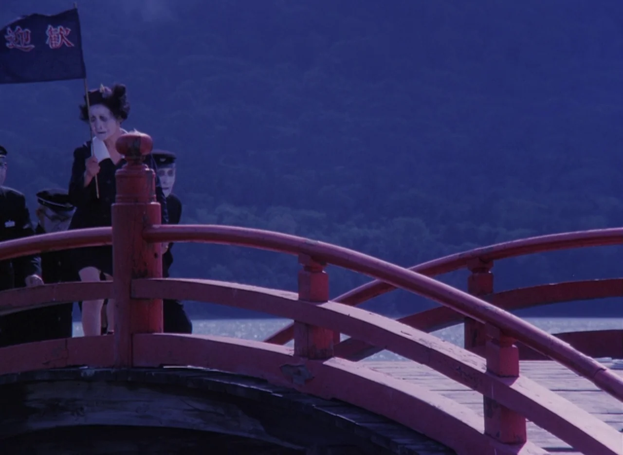 Pastoral To Die In The Country (1974) movie still: medium — A group of people wearing uniforms and white face makeup crosses a red bridge. They move…
