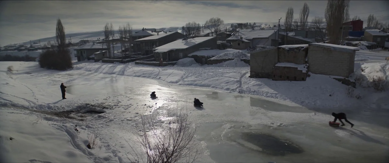 About Dry Grasses (2023) movie still: wide — A snow-covered village borders a wide, icy, frozen pond under an overcast sky. Four individuals move…