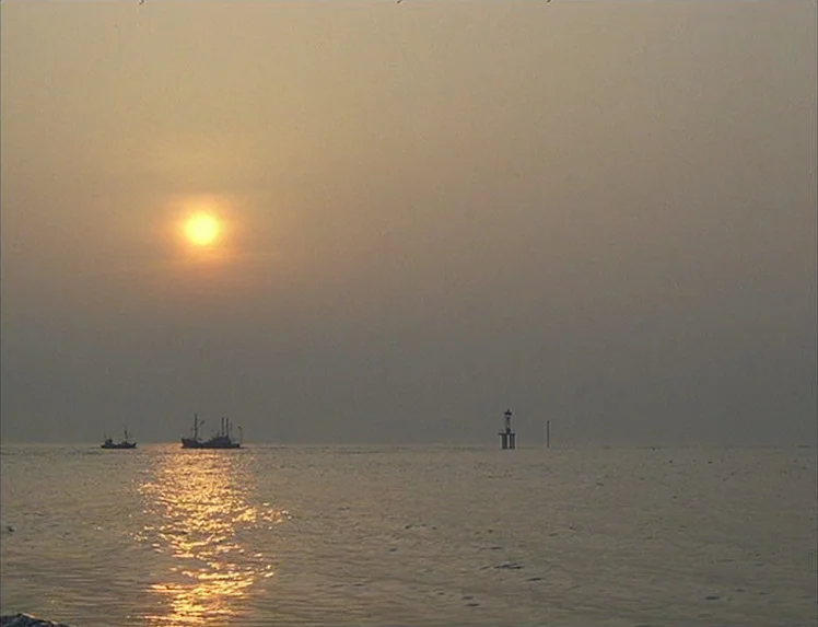 First Name: Carmen (1983) movie still: establishing — A calm expanse of sea under a hazy golden hour sky. Two small fishing vessels sit in the…