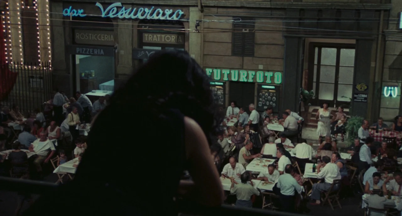 Roma (1972) movie still: medium — A crowded open-air dining area at night features multiple tables with patrons. A woman stands on a balcony in the…