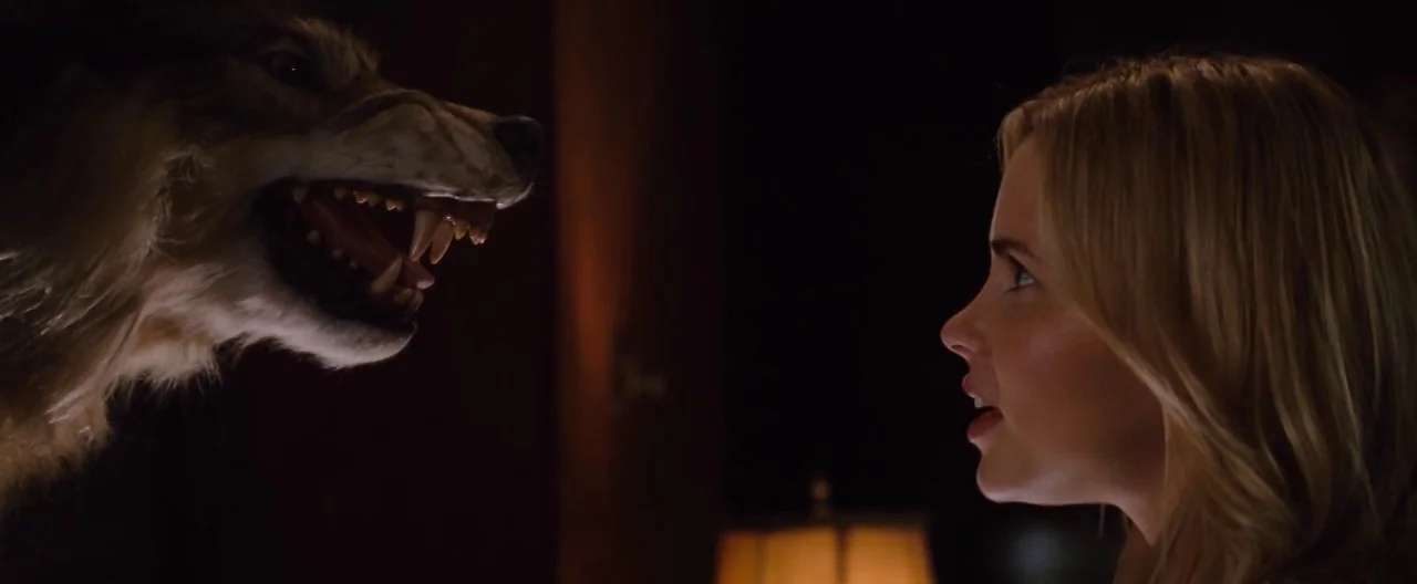 The Cabin In The Woods (2012) movie still: medium — A dark, dimly lit den features a taxidermied wolf head protruding from the wall. A young woman…