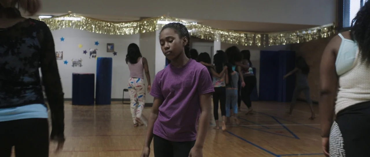 The Fits (2016) movie still: medium — A wooden-floored activity hall is decorated with gold tinsel and small star cutouts. A young girl stands in…
