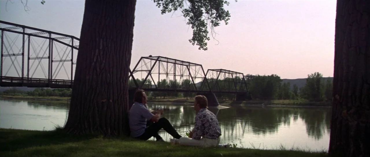 Thunderbolt & Lightfoot (1974) movie still: wide — A river flows beneath a large metal truss bridge in an open rural landscape. Two men sit on the…